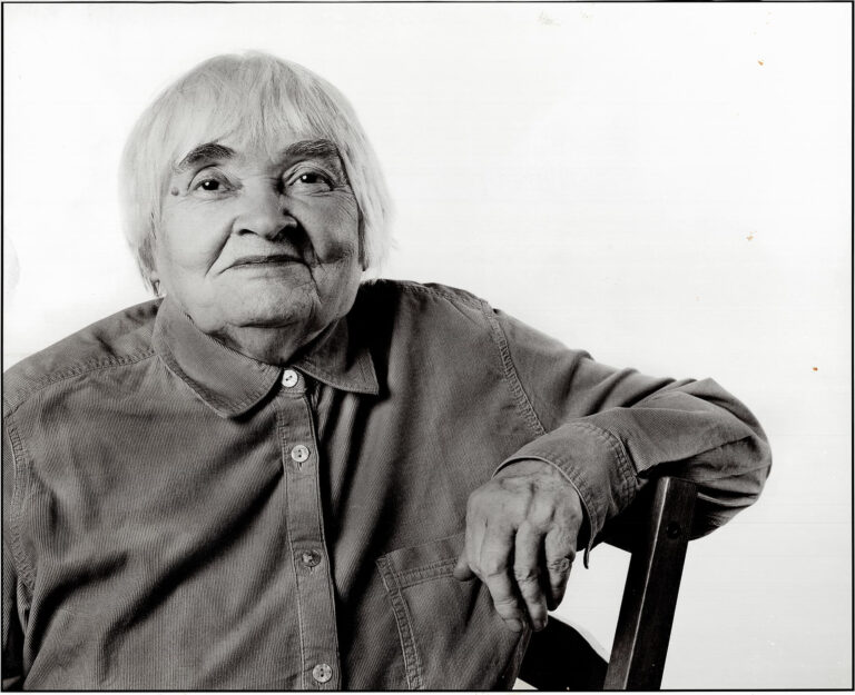 Starsza kobieta siedząca na krześle, patrzy na wprost i lekko się uśmiecha