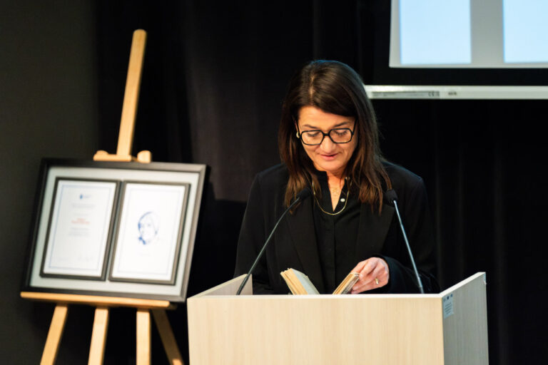 Kobieta w czarnym stroju na mównicy, czytająca jakiś fragment w trzymanej w dłoni książki