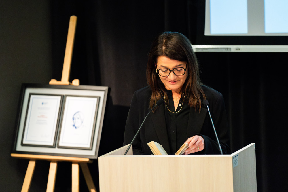 Kobieta w czarnym stroju na mównicy, czytająca jakiś fragment w trzymanej w dłoni książki