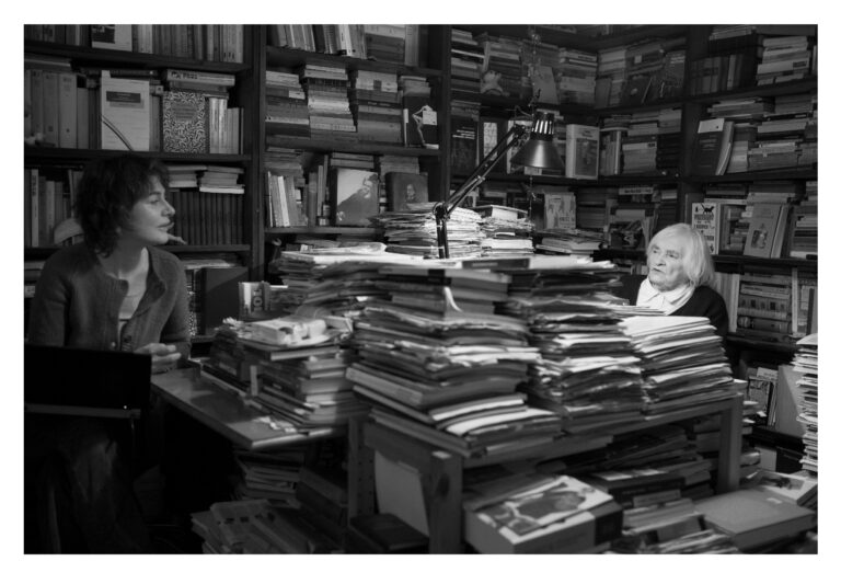 W pokoju zapełnionym regałami z książkami siedzą po obu stronach stołu dwie kobiety, jedna starsza, druga młodsza, rozmawiają ze sobą. Stół założony książkami i dokumentami jest oświetlany przez stojąca pośrodku czarną lampę