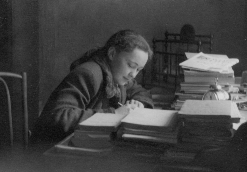 Maria Janion Młoda kobieta siedząca przy stole założonym książkami, coś zapisuje. Widać proste metalowe łóżko i duży budzik