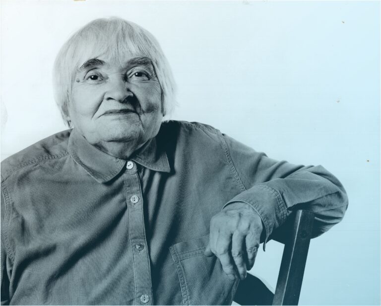 Starsza kobieta siedząca na krześle, patrzy na wprost i lekko się uśmiecha