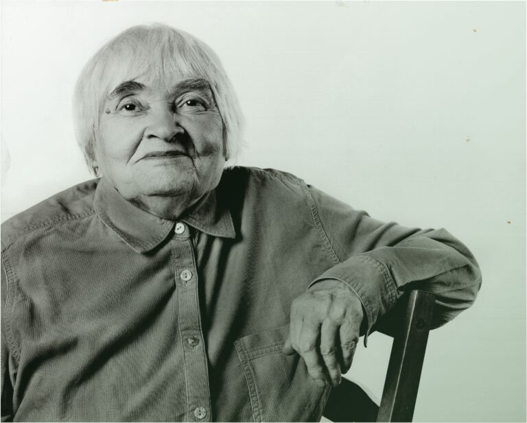 Starsza kobieta siedząca na krześle, patrzy na wprost i lekko się uśmiecha