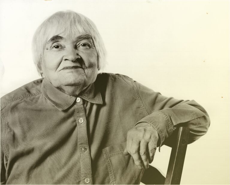 Starsza kobieta siedząca na krześle, patrzy na wprost i lekko się uśmiecha