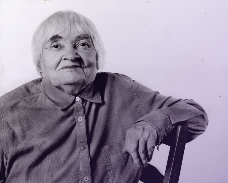 Starsza kobieta siedząca na krześle, patrzy na wprost i lekko się uśmiecha