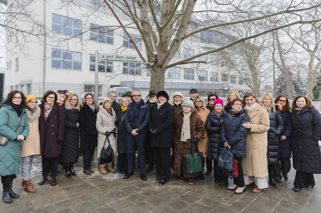 Na pierwszym planie na ulicy grupa ludzi pozująca do zdjęcia, na drugim tabliczka z napisem ul. prof. Marii Janion, dalej widać szaro-biały kilkupiętrowy budynek i kilka drzew pokrytych delikatnie śniegiem